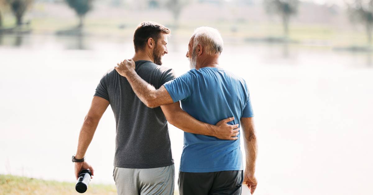 Back view of happy athletic man and his senior father walking embraced while exercising in nature.