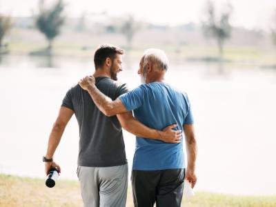 Back view of happy athletic man and his senior father walking embraced while exercising in nature.