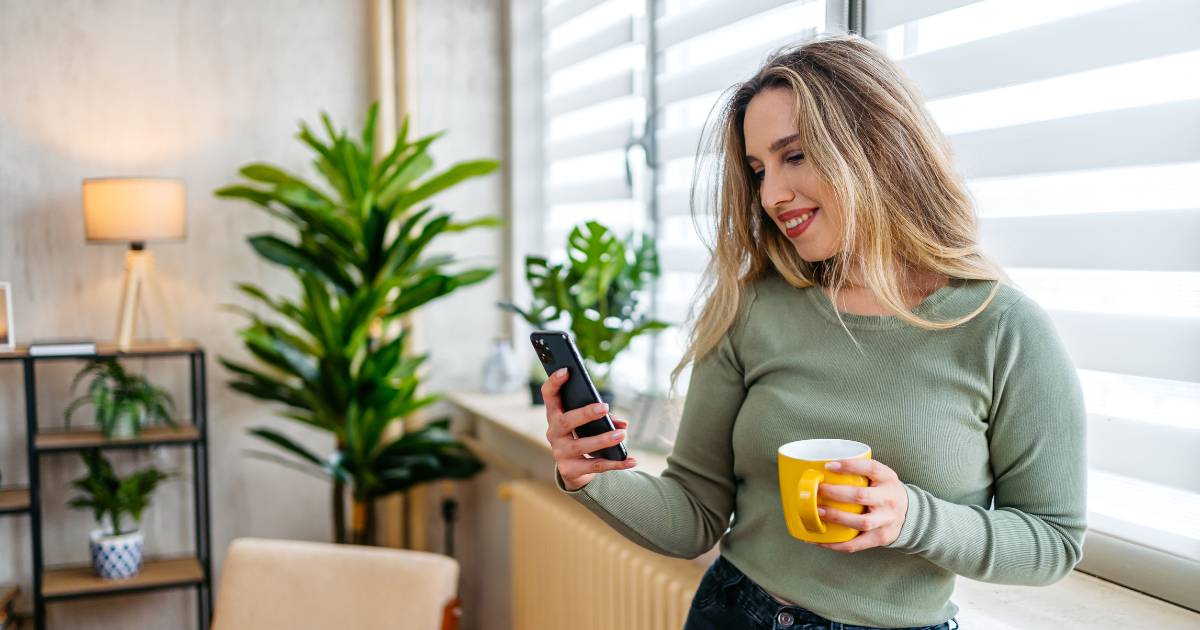 Young Woman Drinking Coffee And Using Phone By The Window At Home