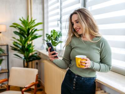 Young Woman Drinking Coffee And Using Phone By The Window At Home
