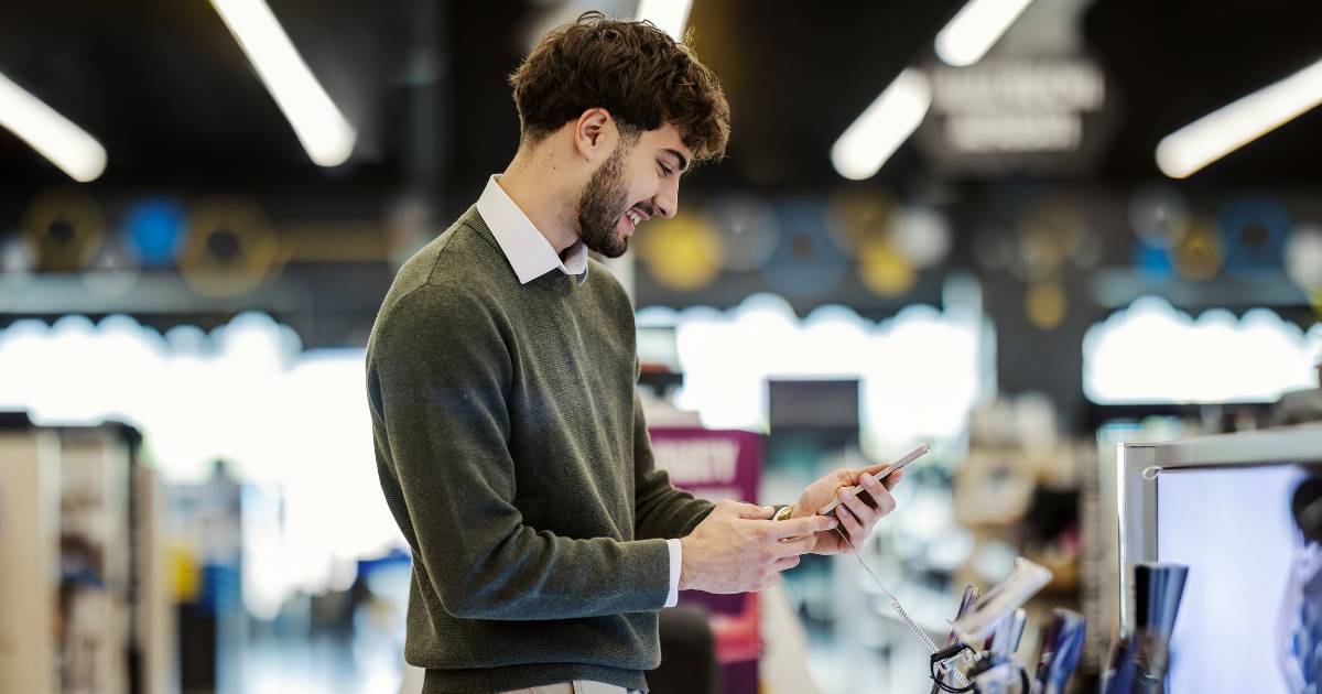 Young adult man smiling while holding and interacting with a smartphone display model, comparing features and considering a purchase in a modern electronics retail store.