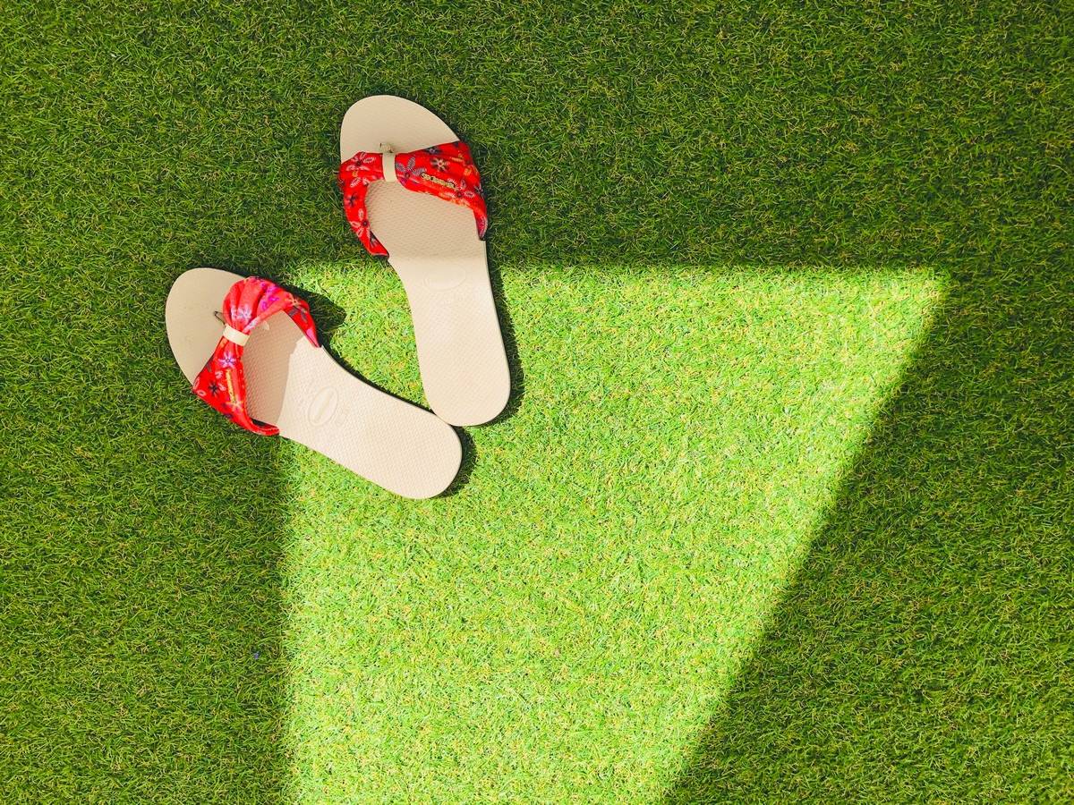 Paire de tongs beige avec des nœuds rouges posées sur une pelouse verte sous une ombre en forme de triangle.