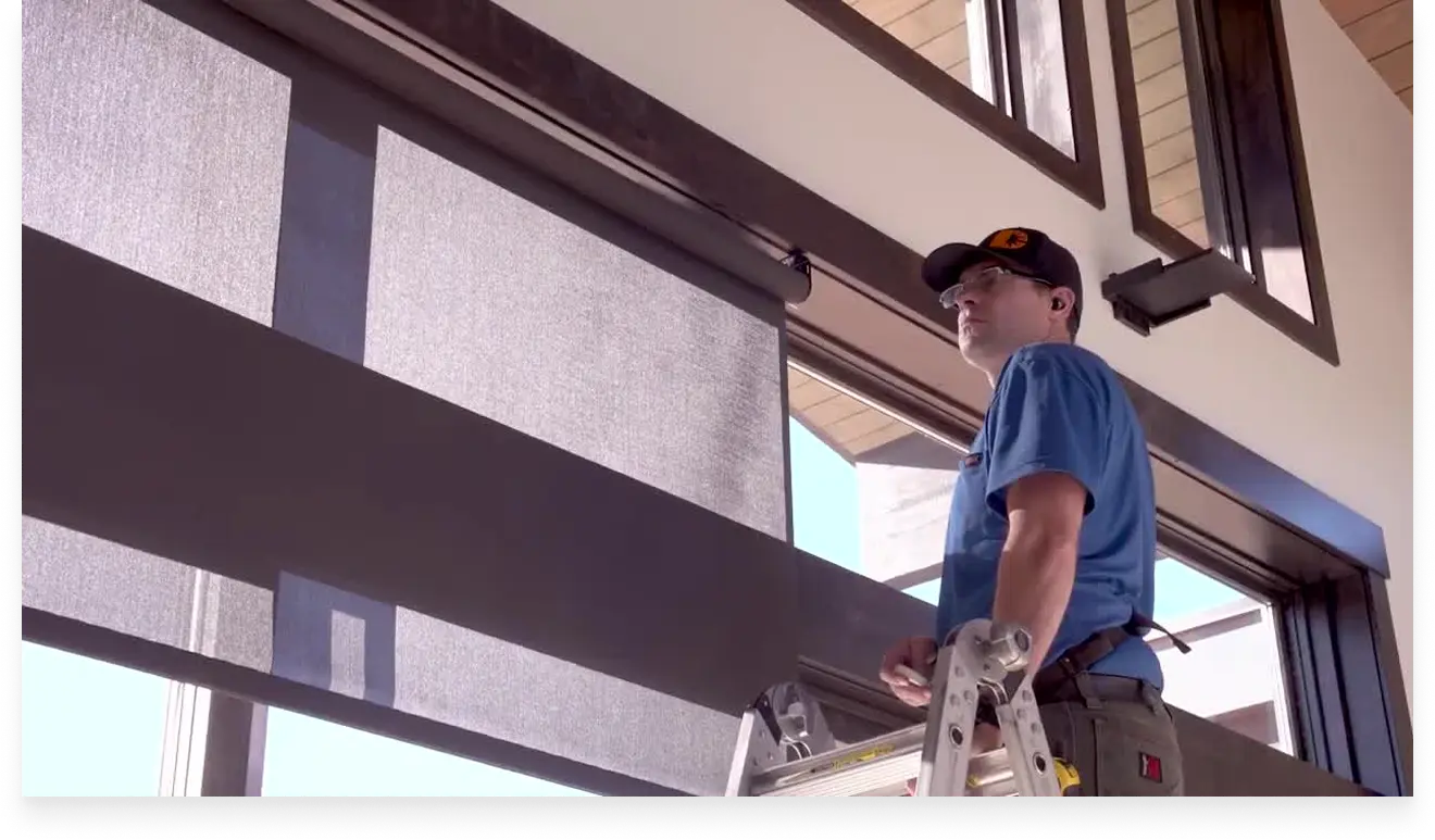 Man in blue shirt and cap on ladder installing or inspecting window blinds in a modern building.