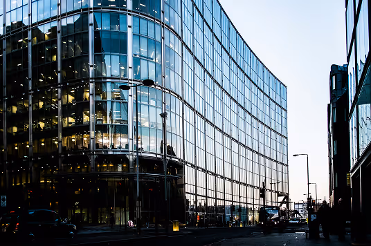 Curved glass office building reflecting the city and sky during dusk with streetlights and people walking nearby.