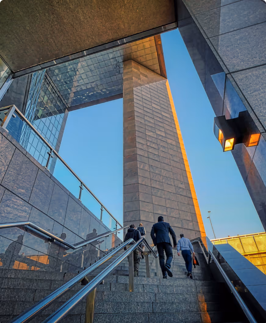 Business people walking up stone stairs toward a modern skyscraper with glass and concrete facades under a clear blue sky.