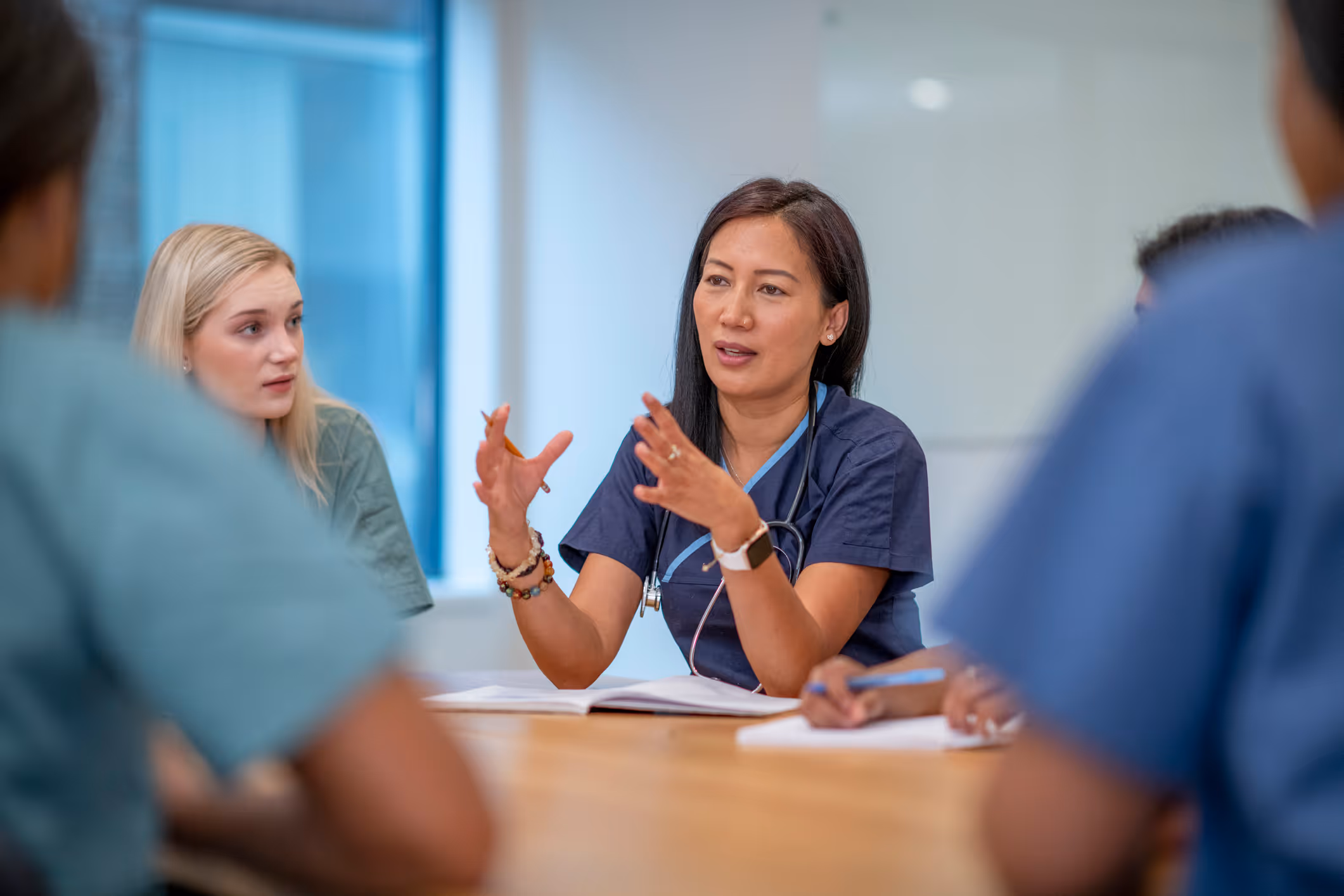 A female medical professional in navy scrubs explaining something to colleagues around a table in a meeting room.