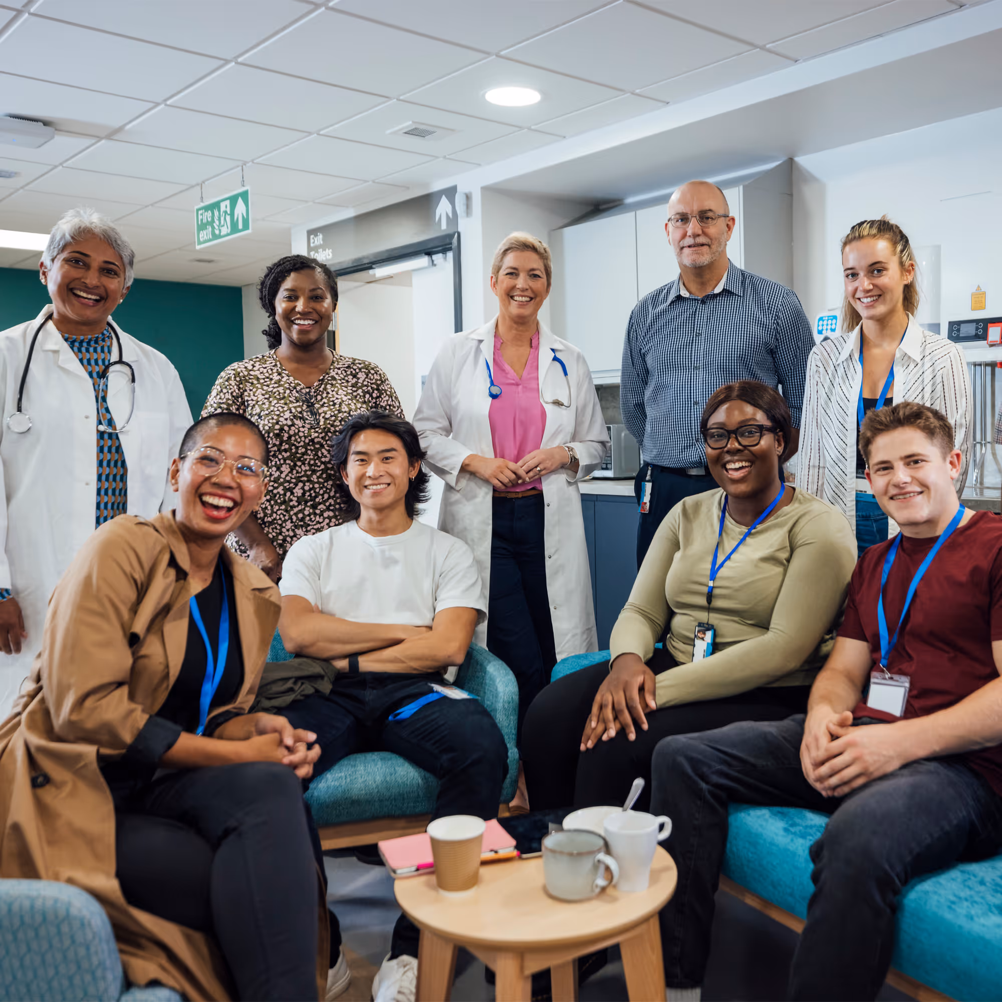A diverse group of nine smiling healthcare professionals and students in a hospital break room with coffee cups on a small table.
