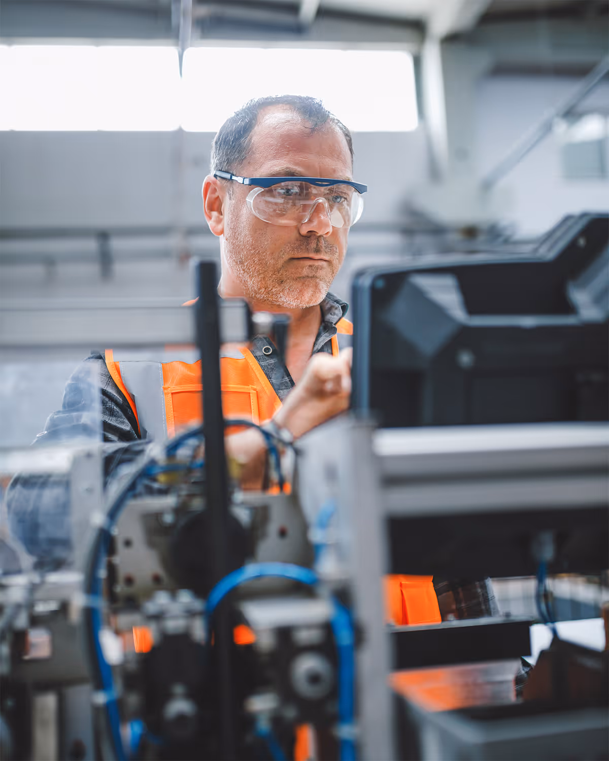 Man wearing safety glasses and an orange vest operating industrial machinery in a factory.