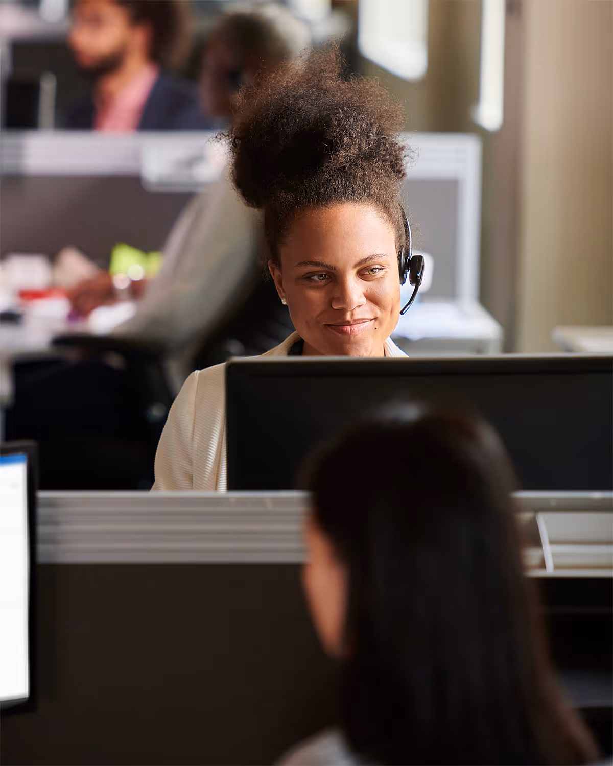 Smiling woman with curly hair wearing a headset, working at a computer in a busy office.