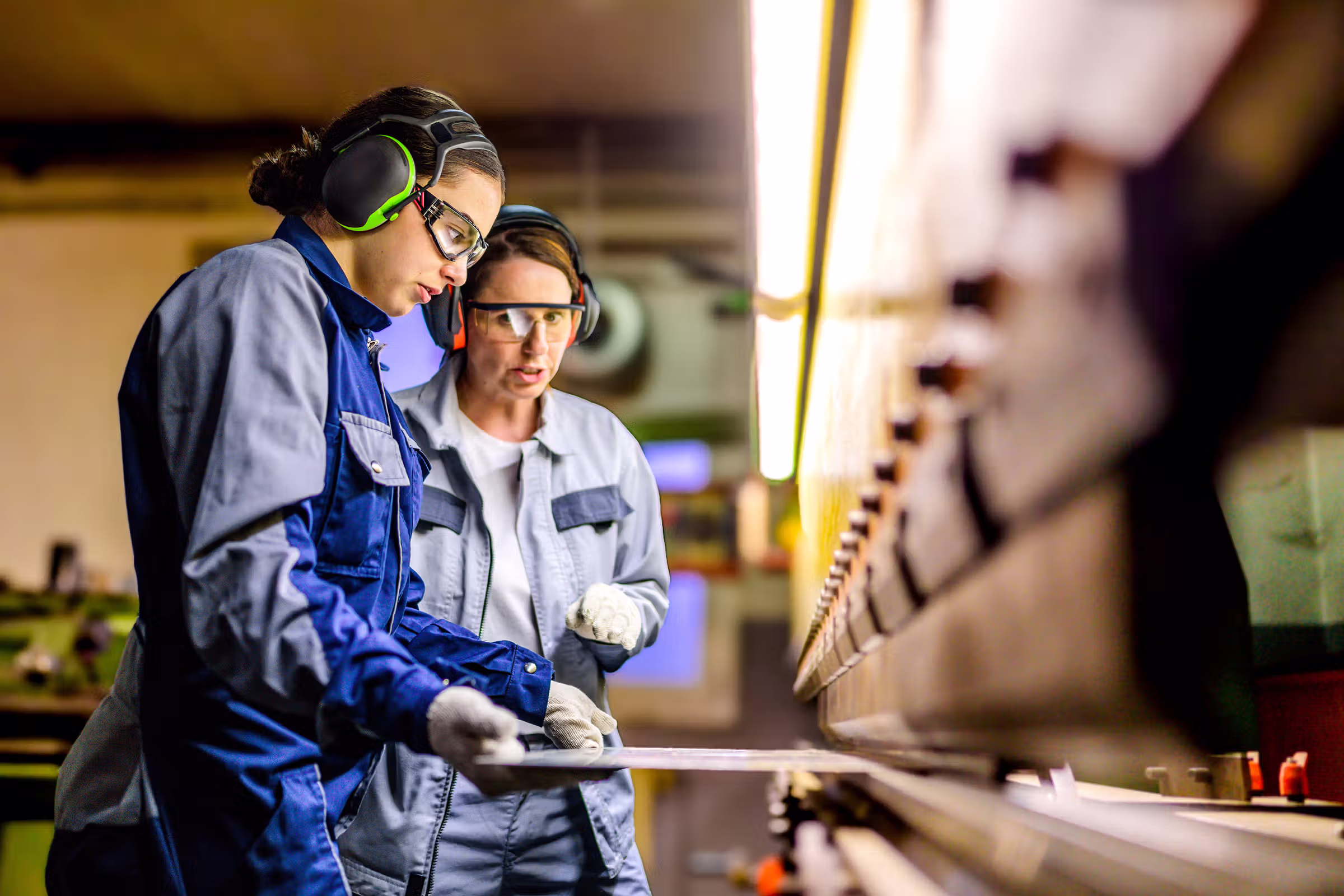 Two female factory workers wearing safety gear operate a large industrial machine together.