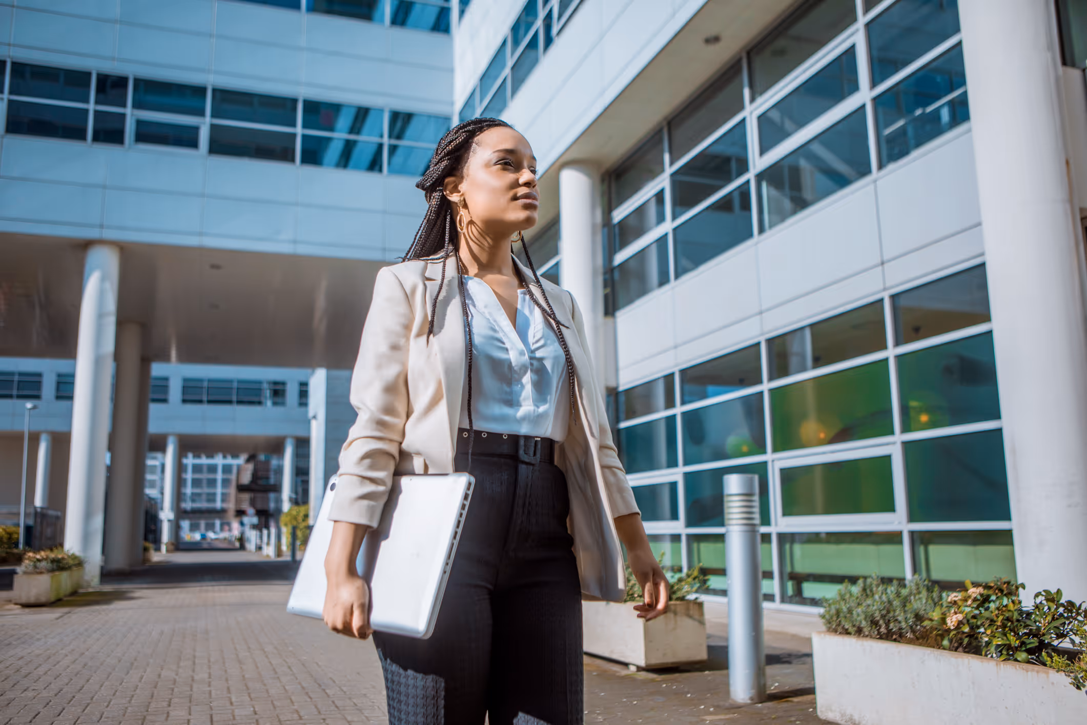Confident businesswoman walking outdoors holding a laptop with modern office building in the background.