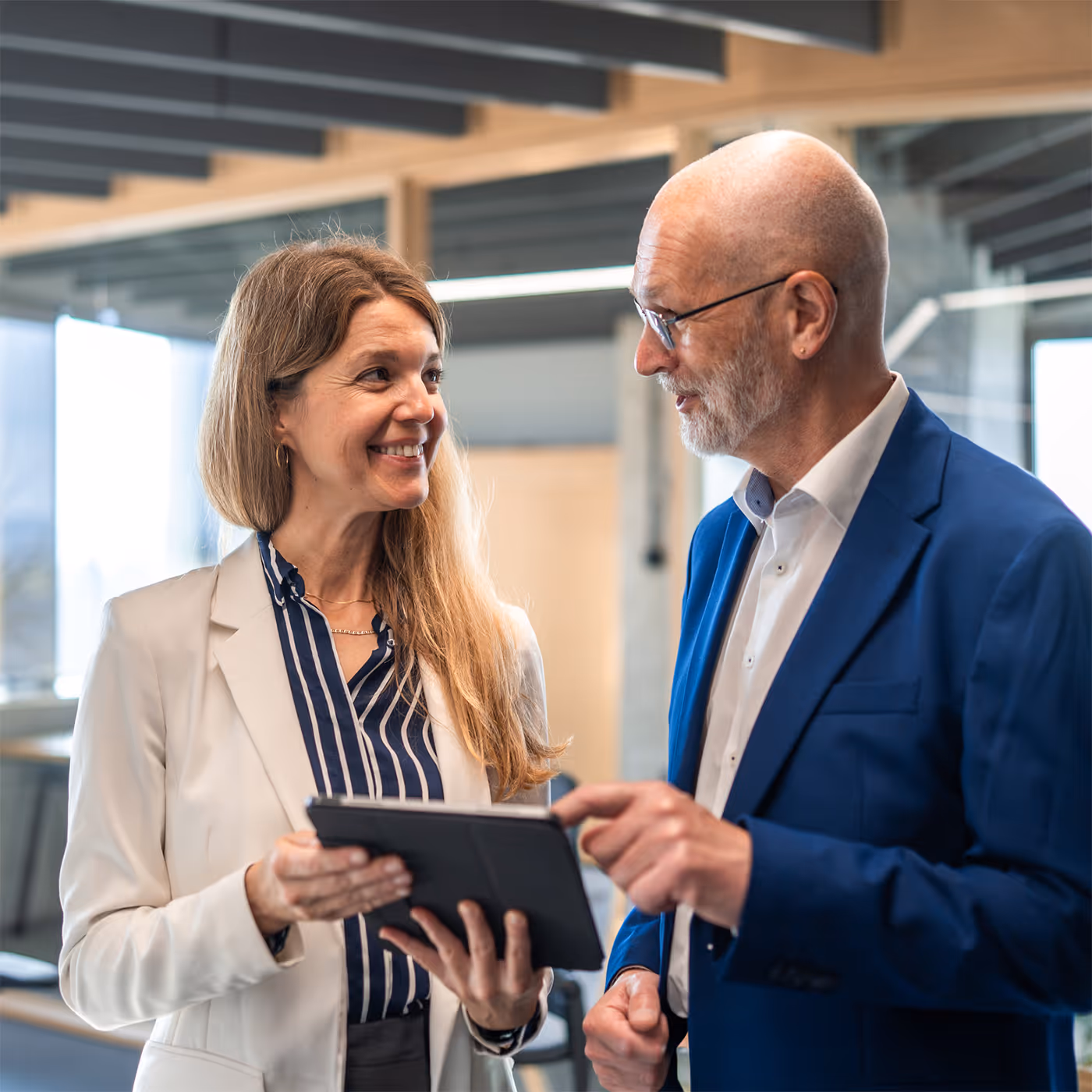 Smiling woman in a white blazer holding a tablet talking to a man in a blue suit in a modern office.