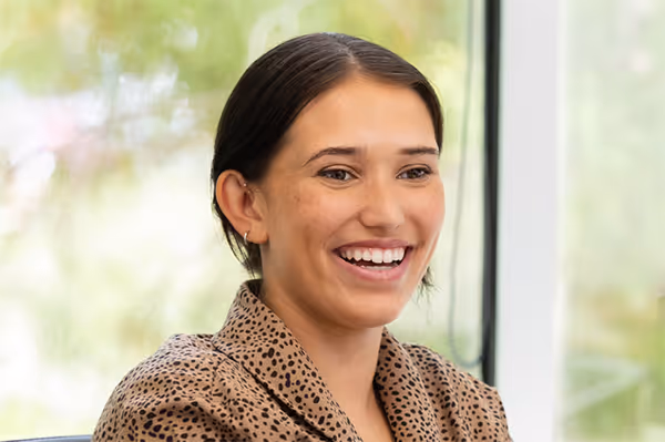 Smiling woman with dark hair wearing a patterned blazer in a bright room with large windows.
