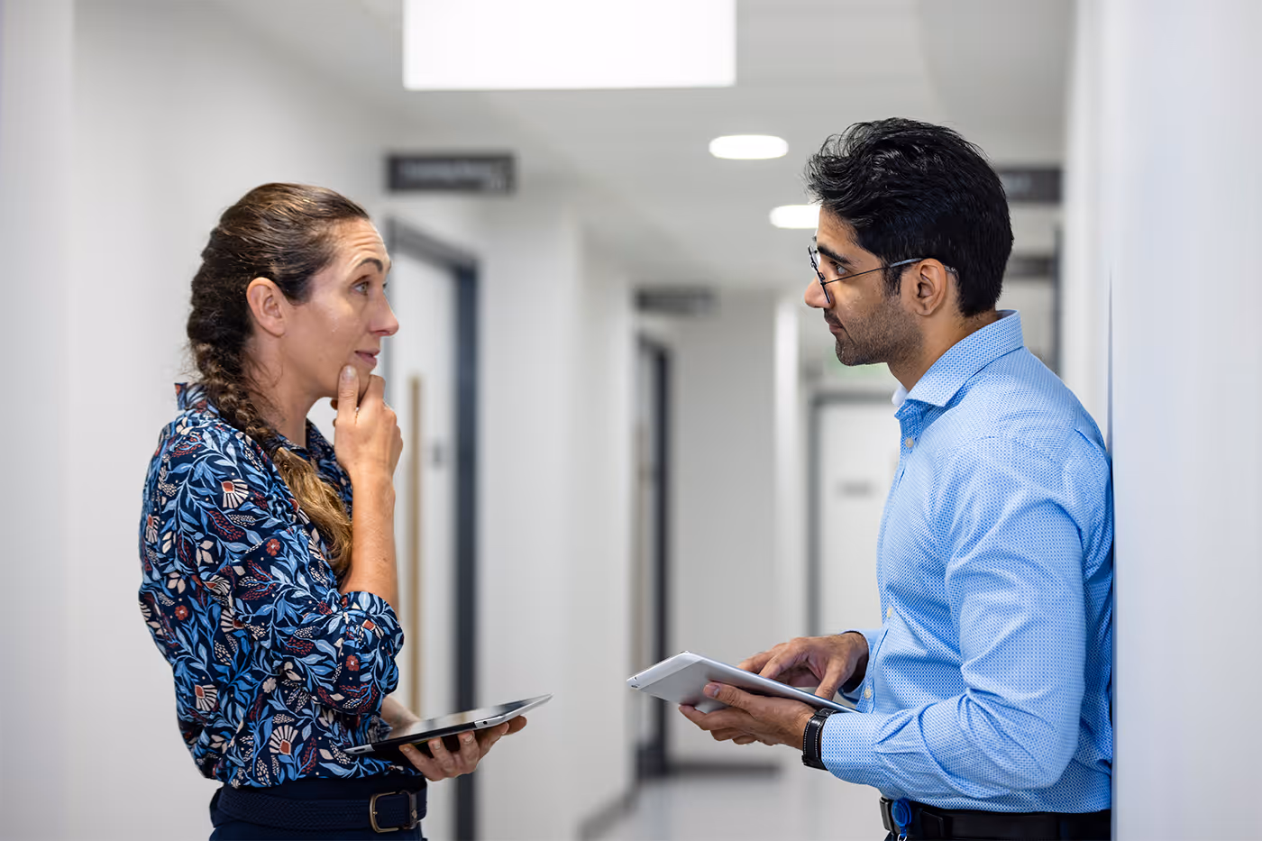 Man and woman having a discussion in a hospital corridor, both holding tablets.