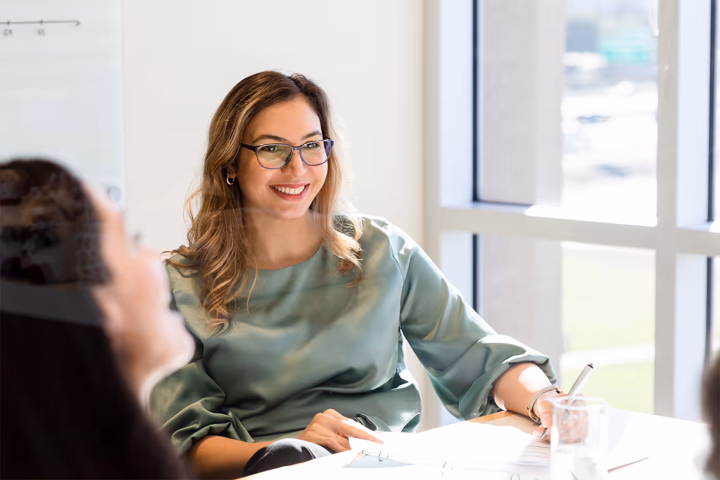 Smiling woman wearing glasses and a green blouse, sitting at a table with papers and a glass of water in a bright office.