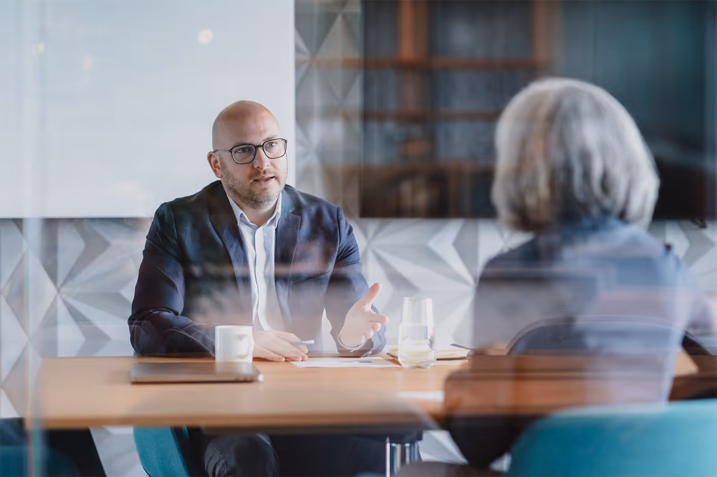 A man in glasses and a blazer engaged in a discussion with a woman across a conference table.