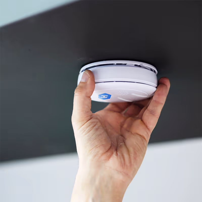 Hand installing or pressing a white smoke detector with a blue hexagon logo on a dark ceiling.