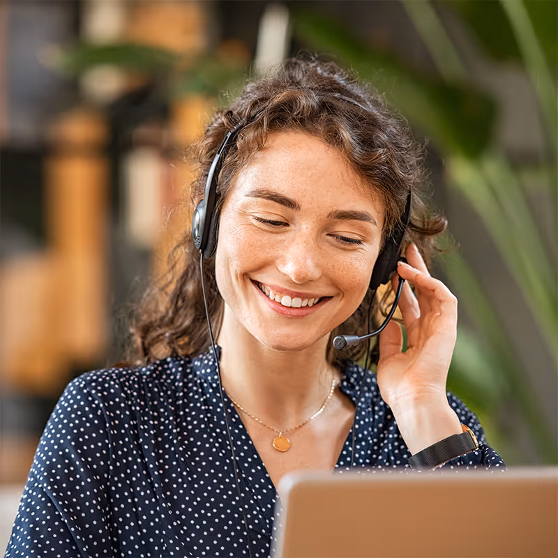 Smiling woman with curly hair wearing a headset and polka dot blouse working on a laptop in a bright room.