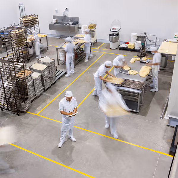Bakery workers in white uniforms preparing dough and placing trays in racks inside a large industrial kitchen.
