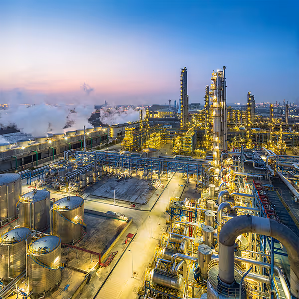 Industrial refinery complex at sunset with illuminated towers, tanks, and pipelines emitting steam.