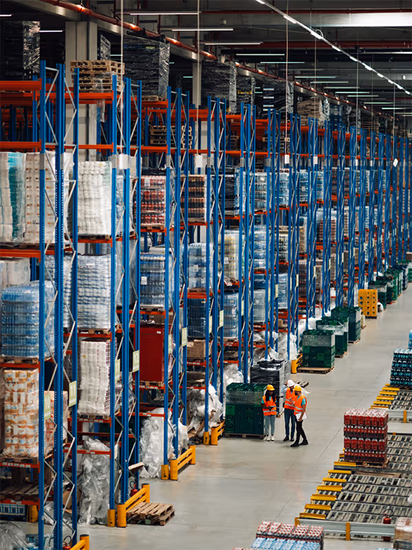 Inside a large warehouse with tall blue and orange storage racks filled with pallets, three workers in orange safety vests and helmets standing on the floor.