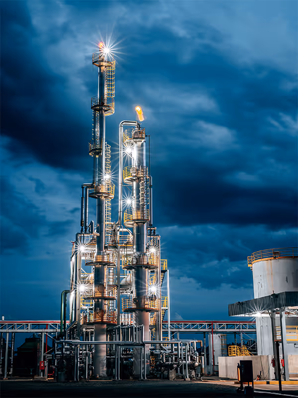Illuminated industrial refinery towers with pipes and platforms against a dark cloudy sky at night.