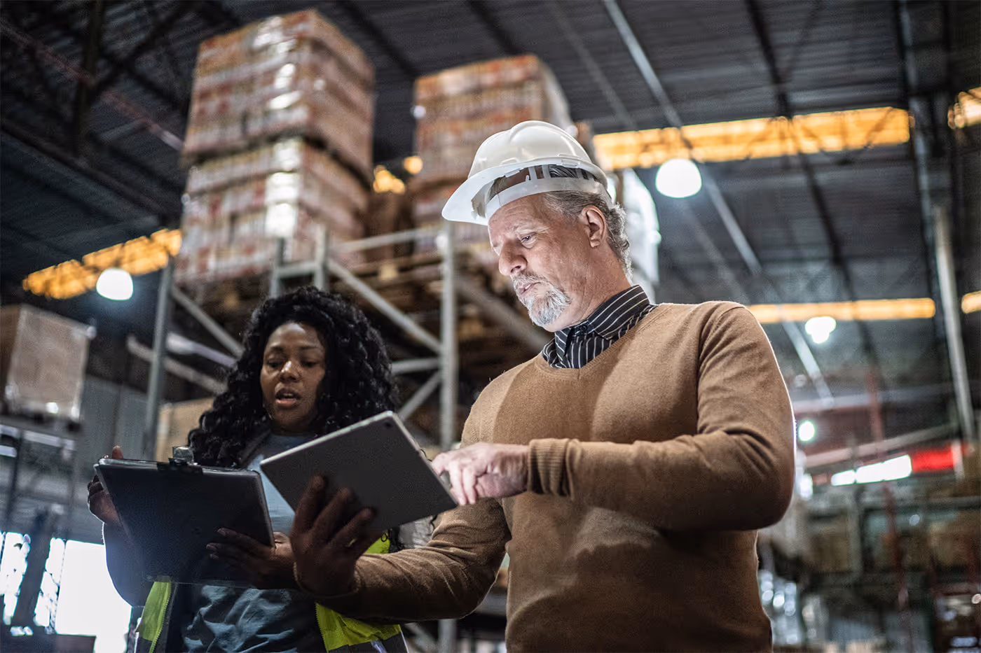Two warehouse workers reviewing information on a tablet and clipboard inside a storage facility with stacked pallets in the background.