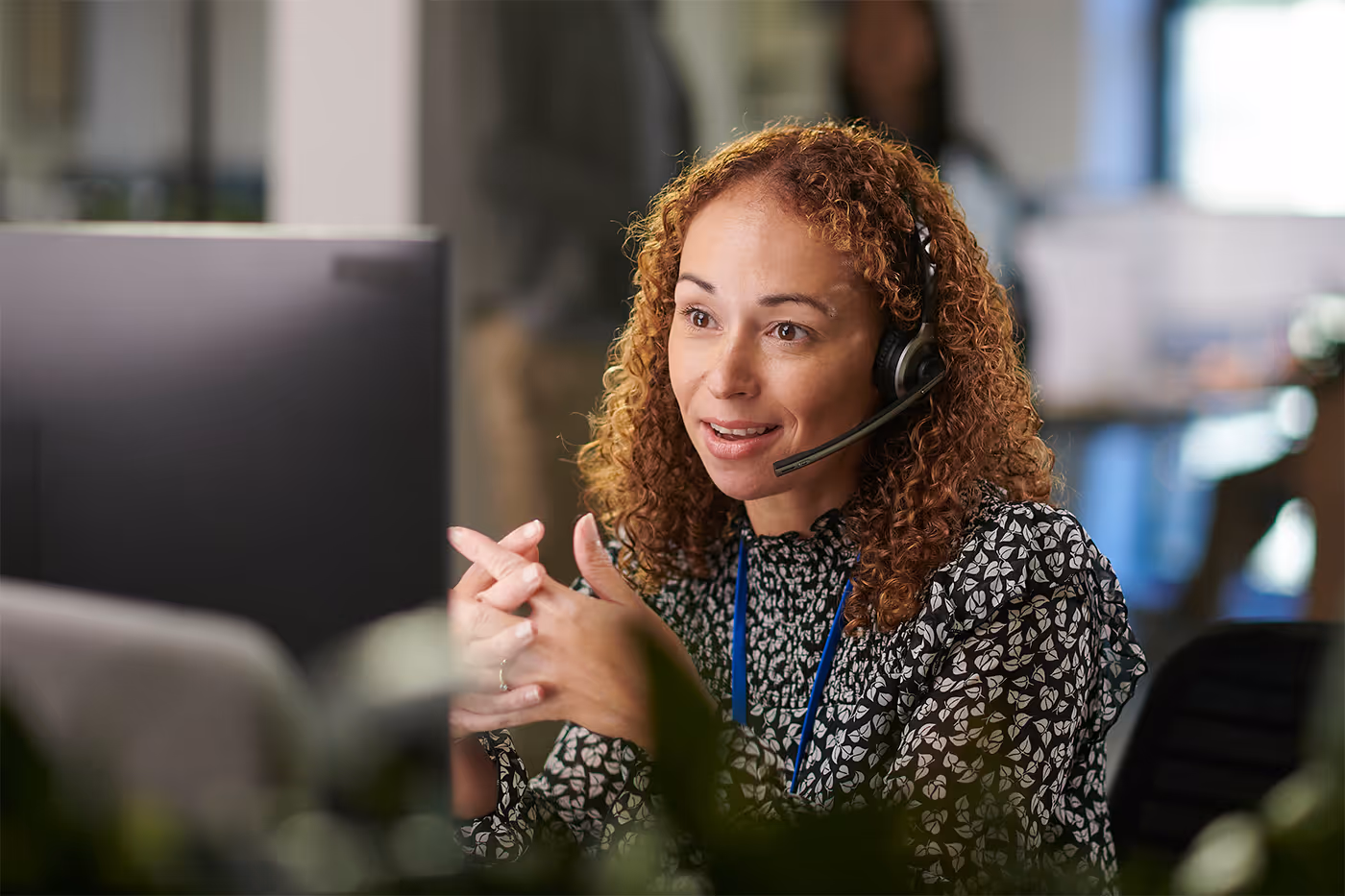 Customer service representative with curly hair wearing a headset and patterned blouse, speaking and gesturing in an office.