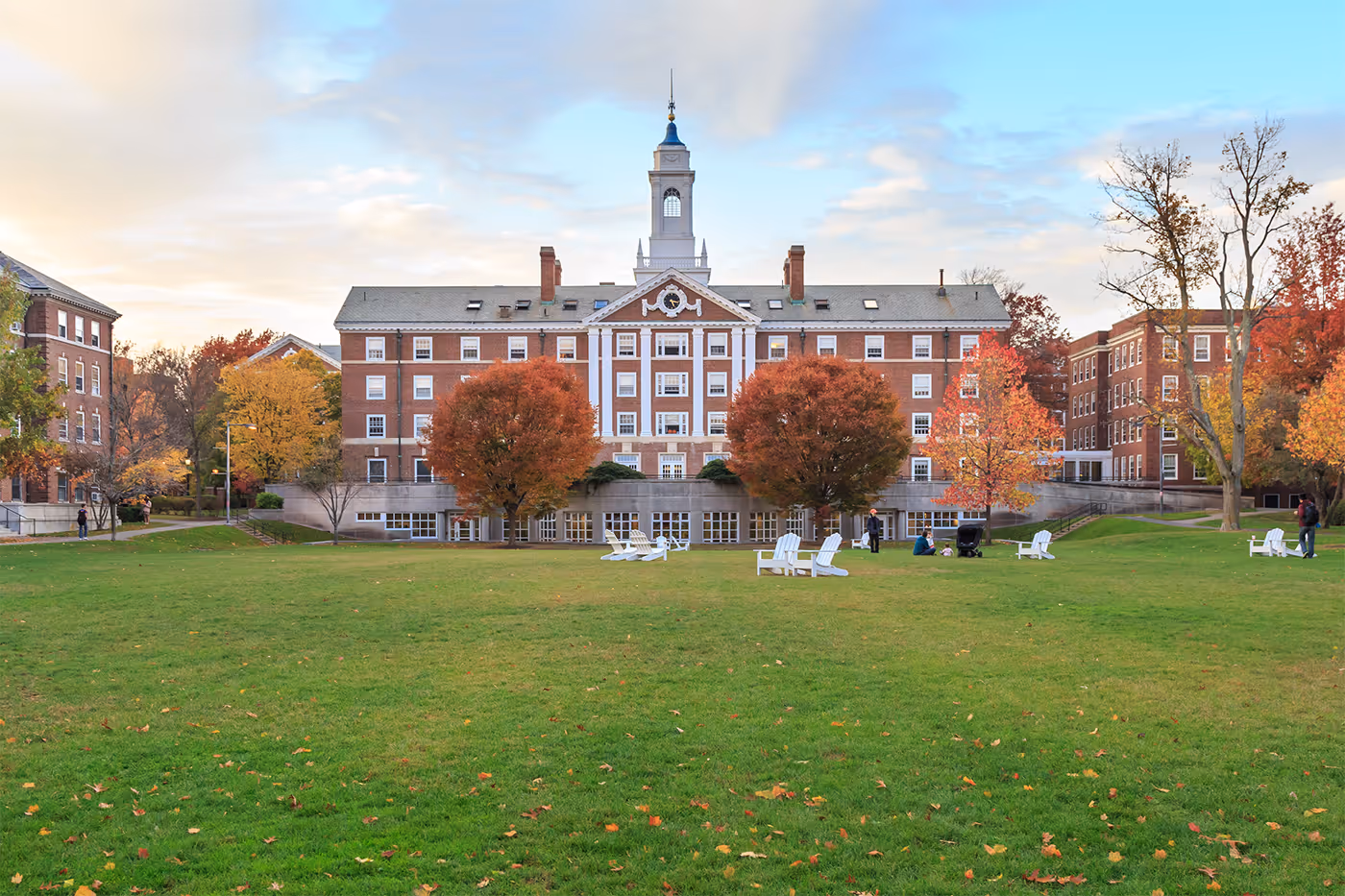 Large red-brick academic building with a central clock tower, surrounded by autumn trees and a grassy lawn with white chairs and a few people.