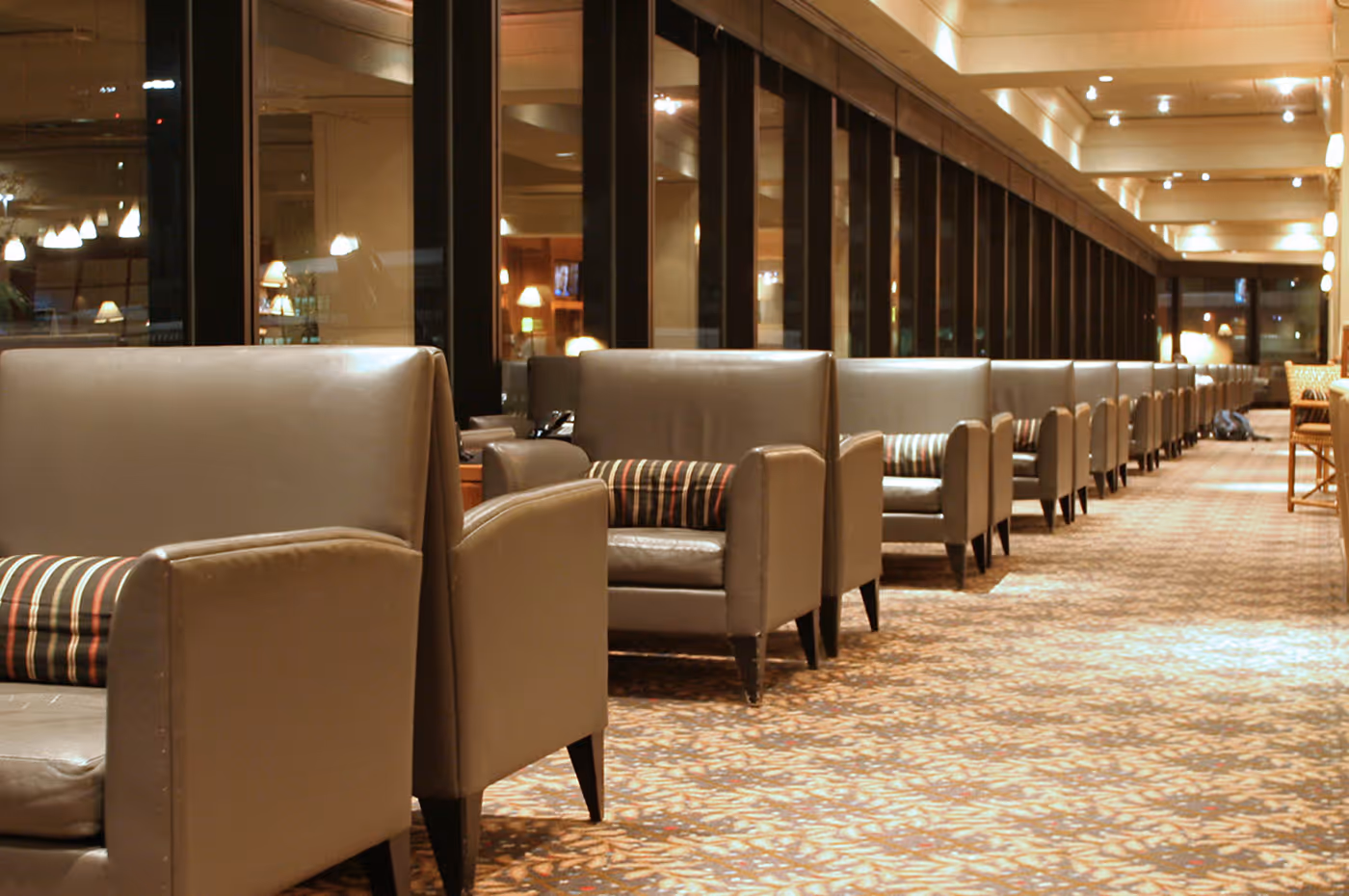 Row of beige leather armchairs with striped cushions in a well-lit hotel lounge with large windows and patterned carpet.