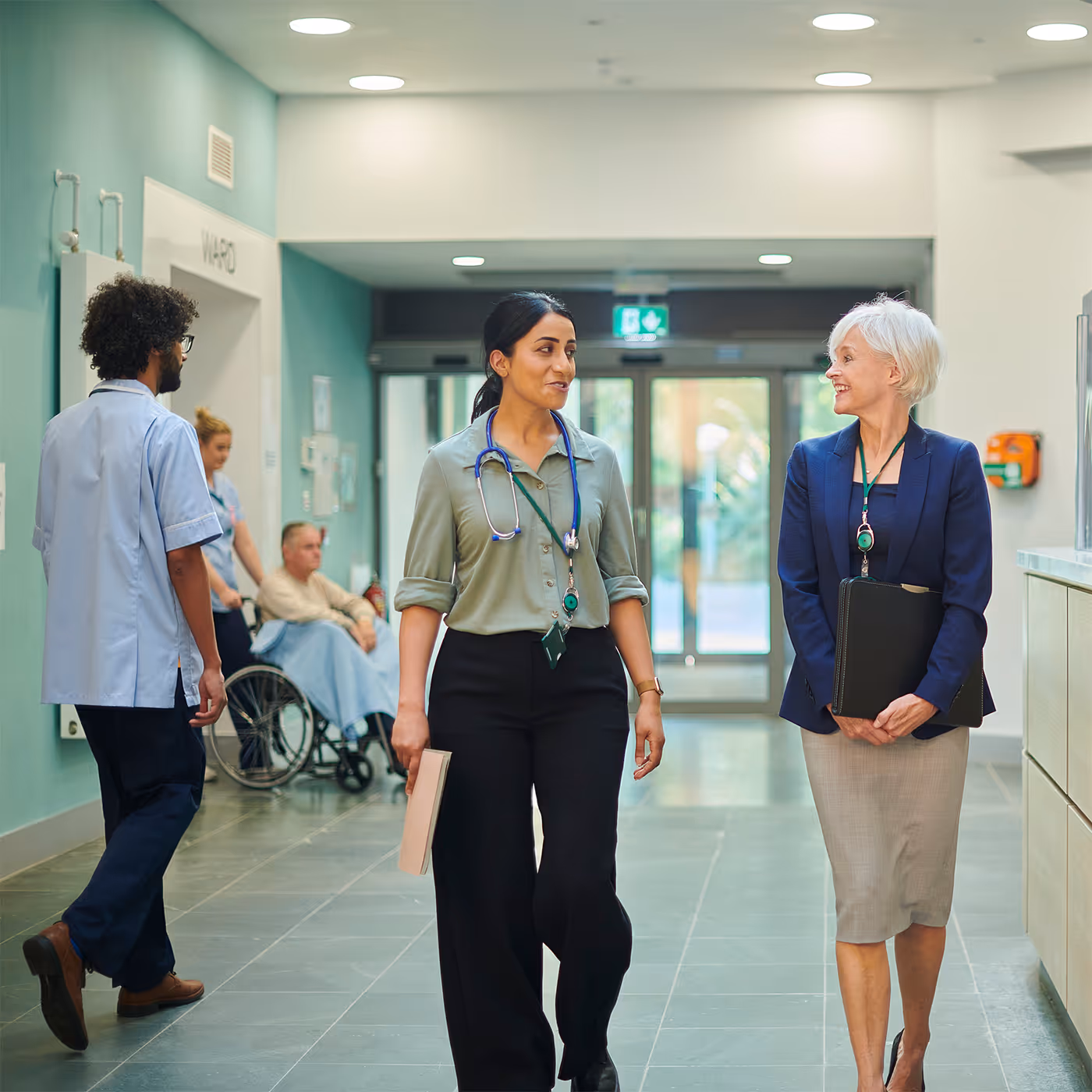 Two healthcare professionals walking and talking in a hospital corridor with a patient in a wheelchair in the background.