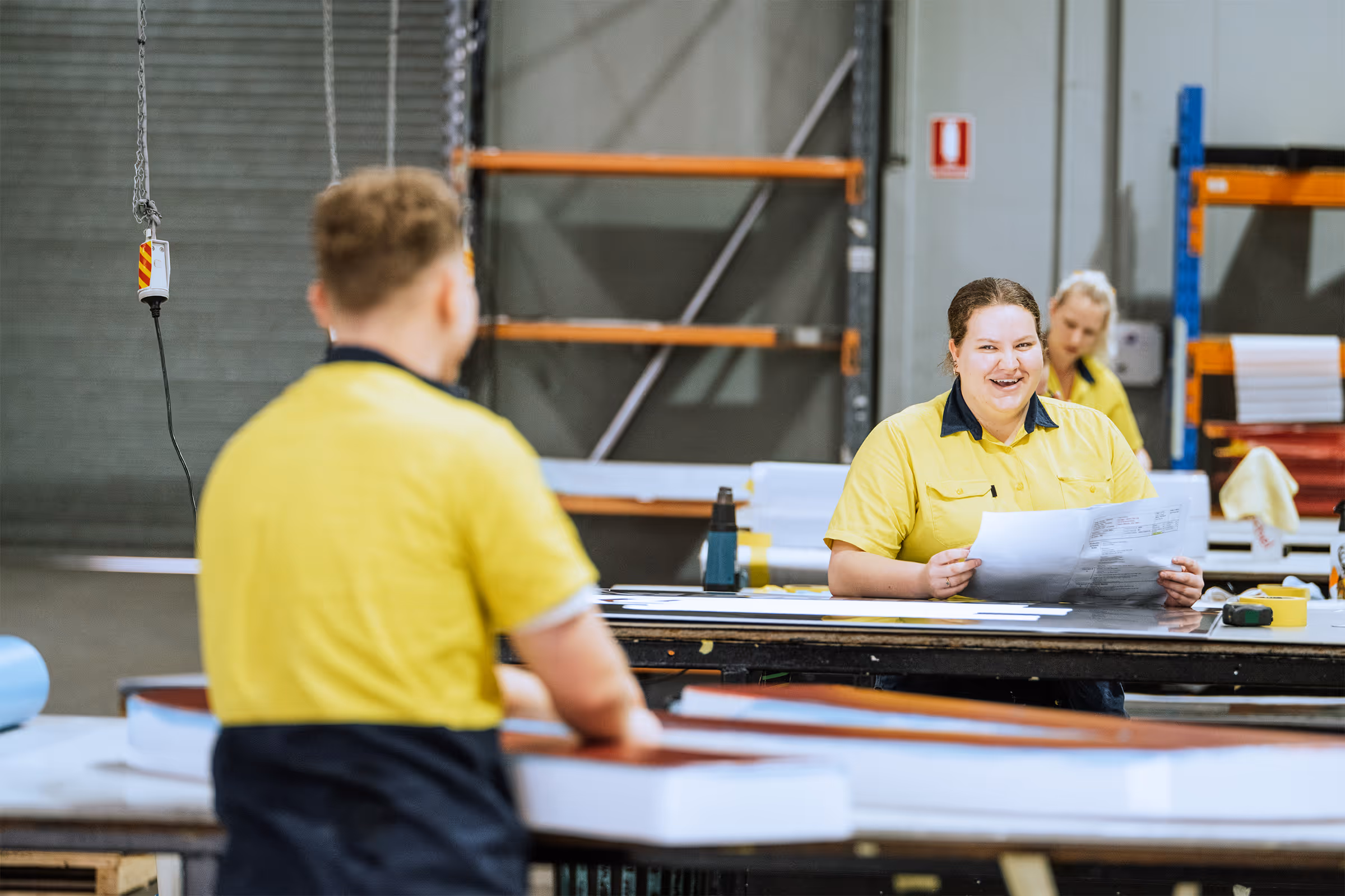Workers in yellow shirts interacting and reviewing papers inside a warehouse or workshop.