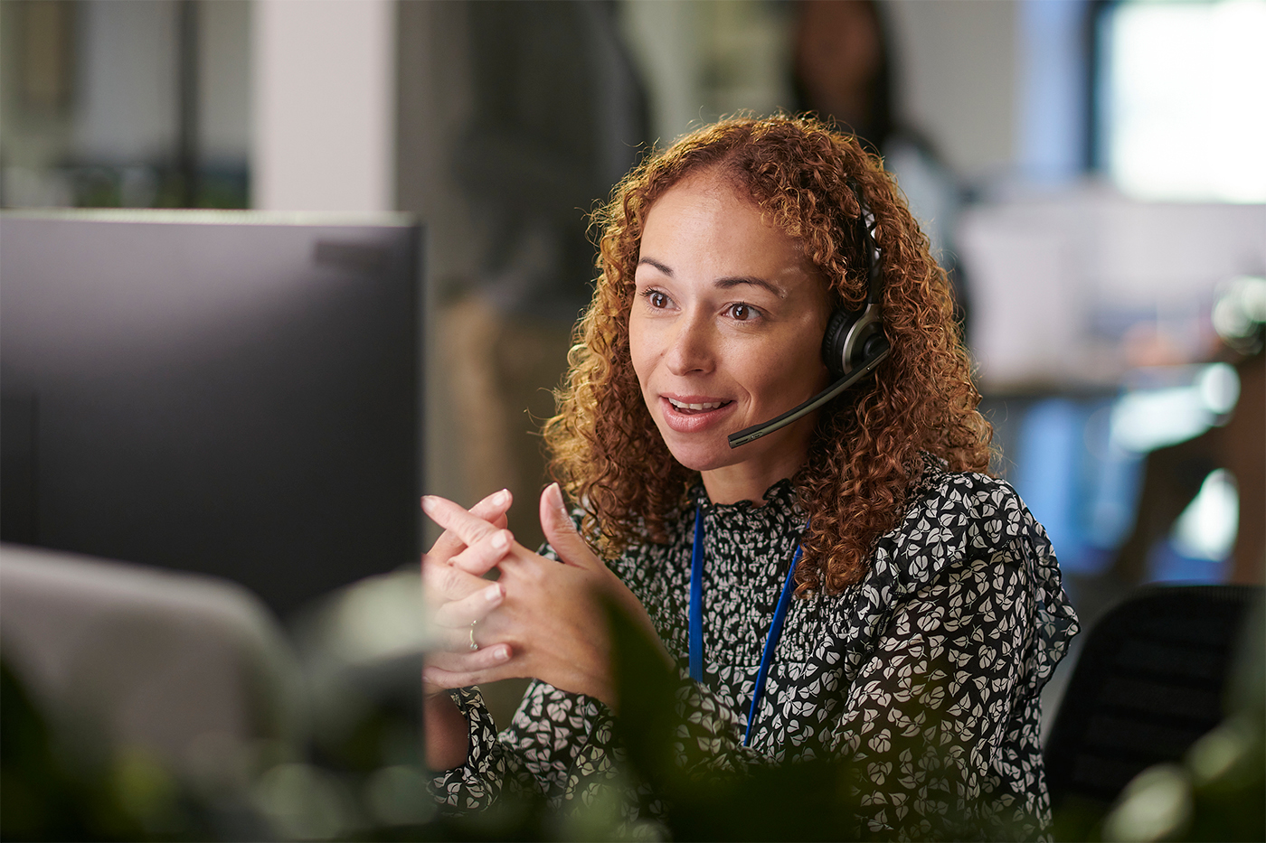 Woman with curly hair wearing a headset and patterned blouse speaking while working at a computer in an office.