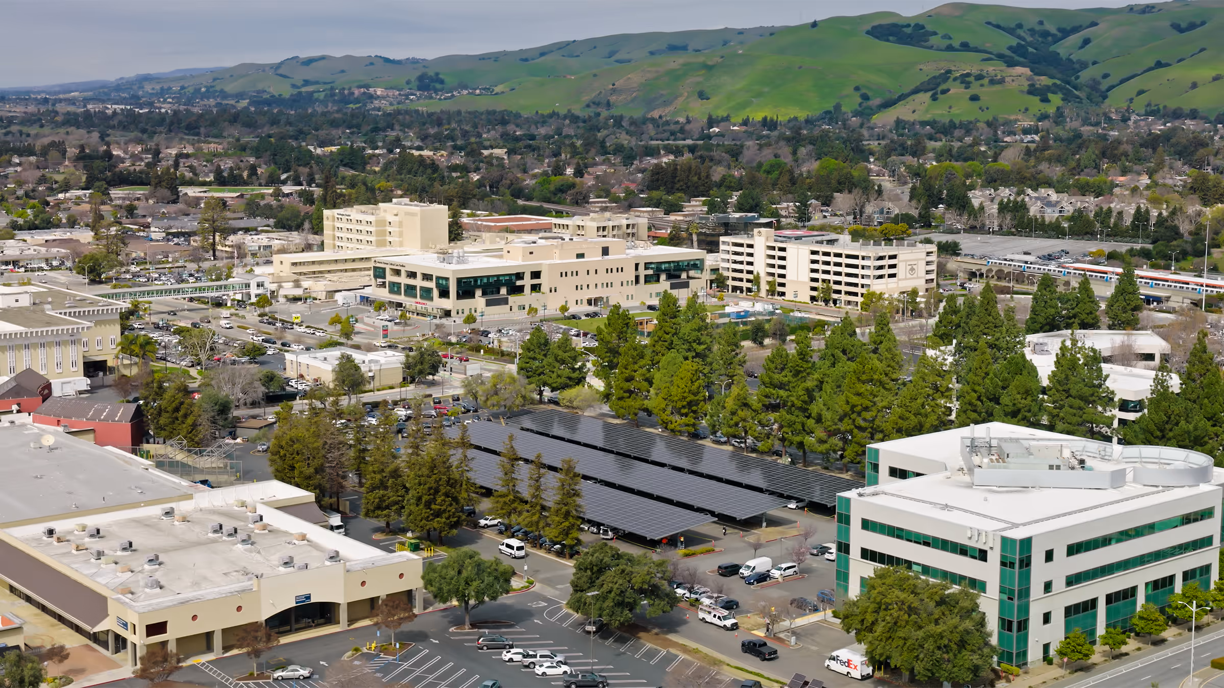 Aerial view of a hospital complex with multiple buildings, parking lots with solar panel canopies, surrounded by green hills and residential areas.