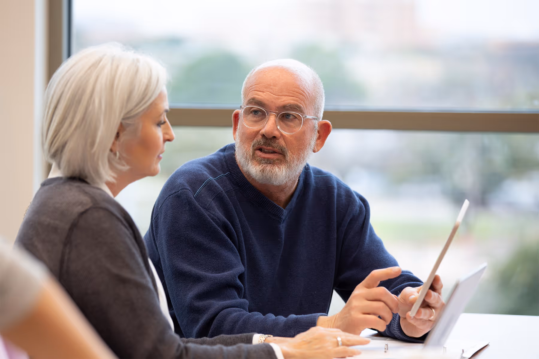 Older man with glasses showing a tablet to an older woman near a large window.