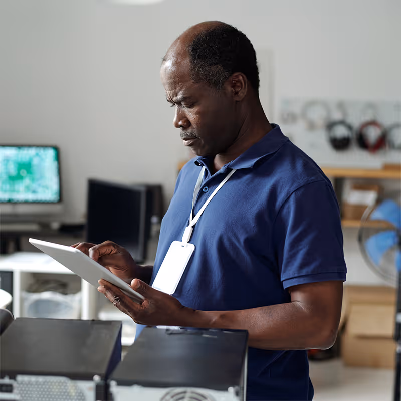 Man with ID badge using a tablet in an office with computers and shelves in background.