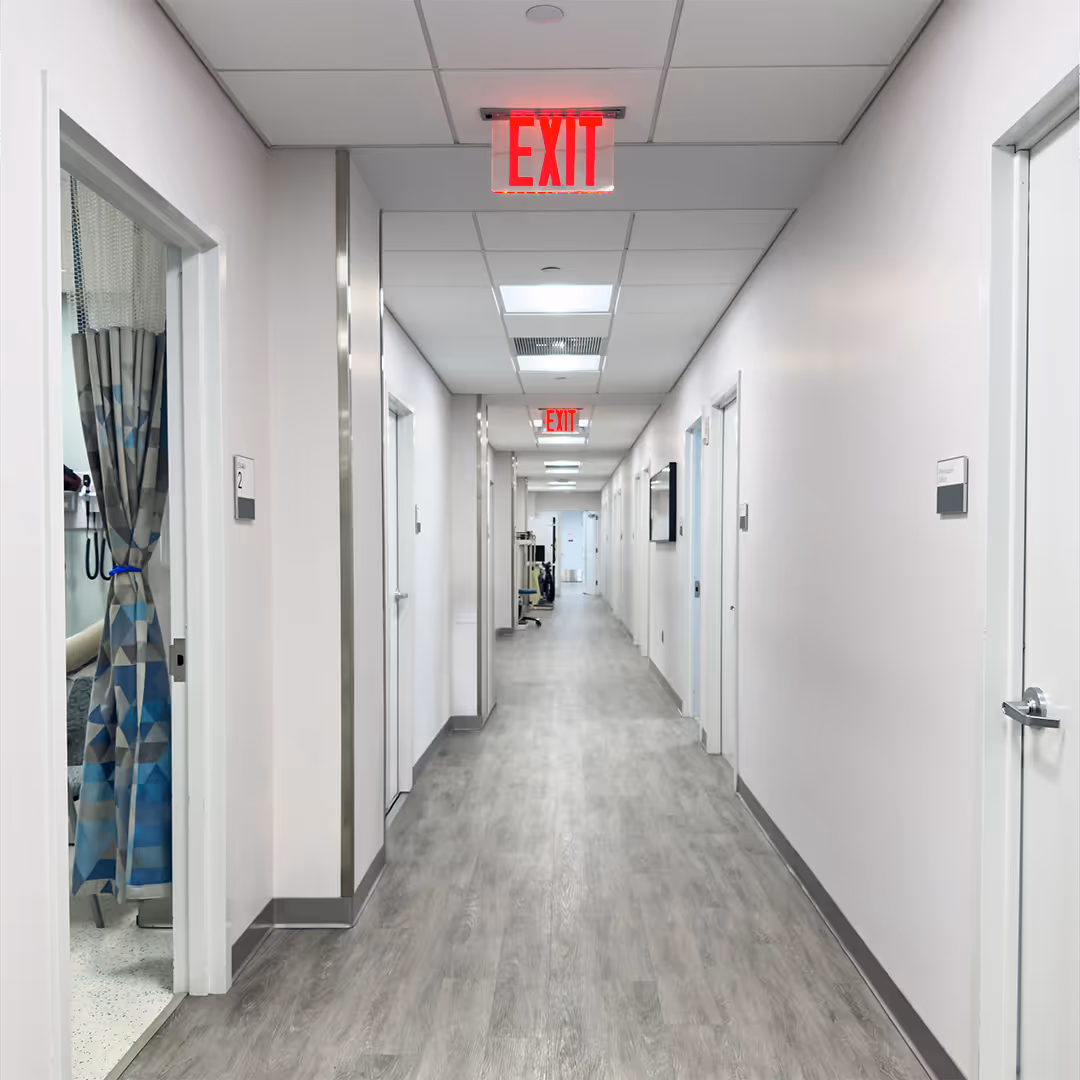 Long hospital hallway with white walls, doors on both sides, and red exit signs on ceiling.