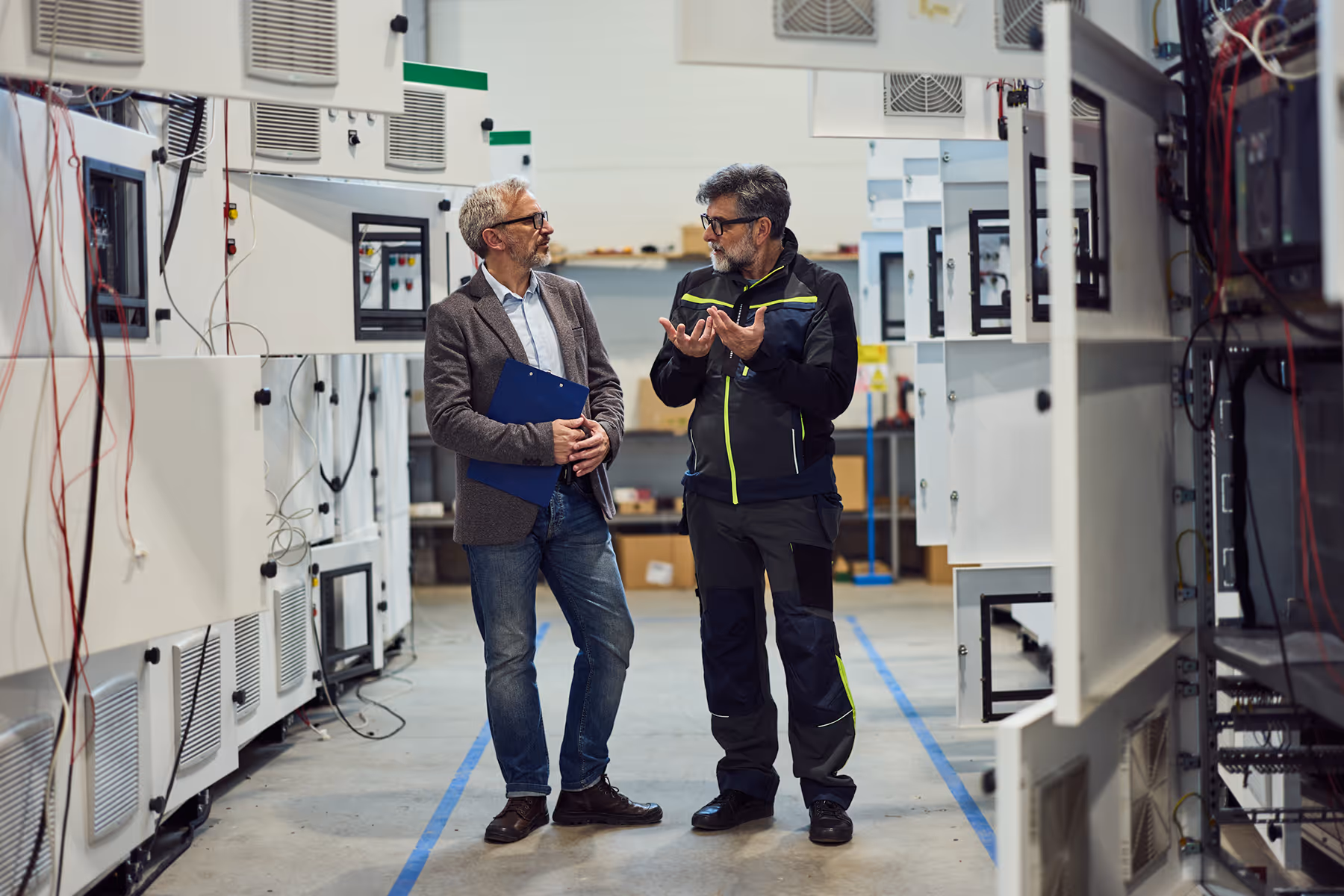 Two men in a factory aisle discussing work surrounded by electrical panels and equipment.