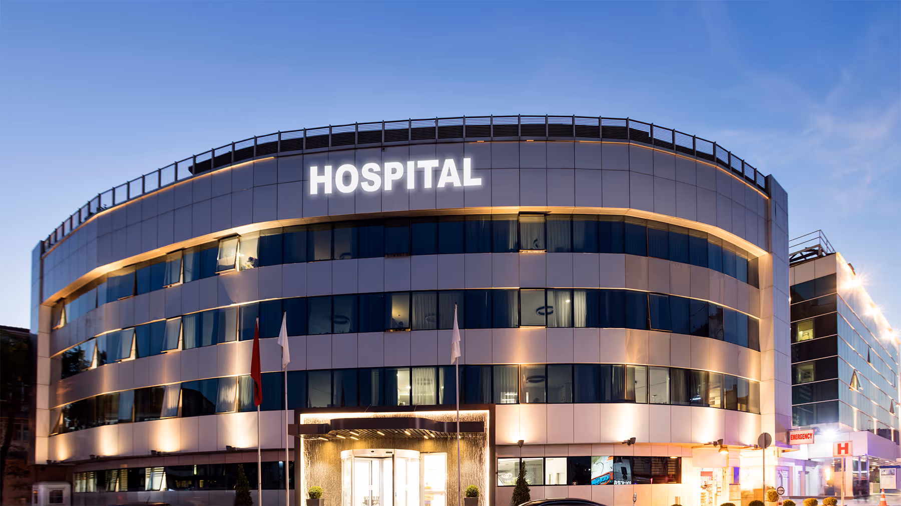 Modern hospital building with illuminated sign and entrance at dusk under clear sky.