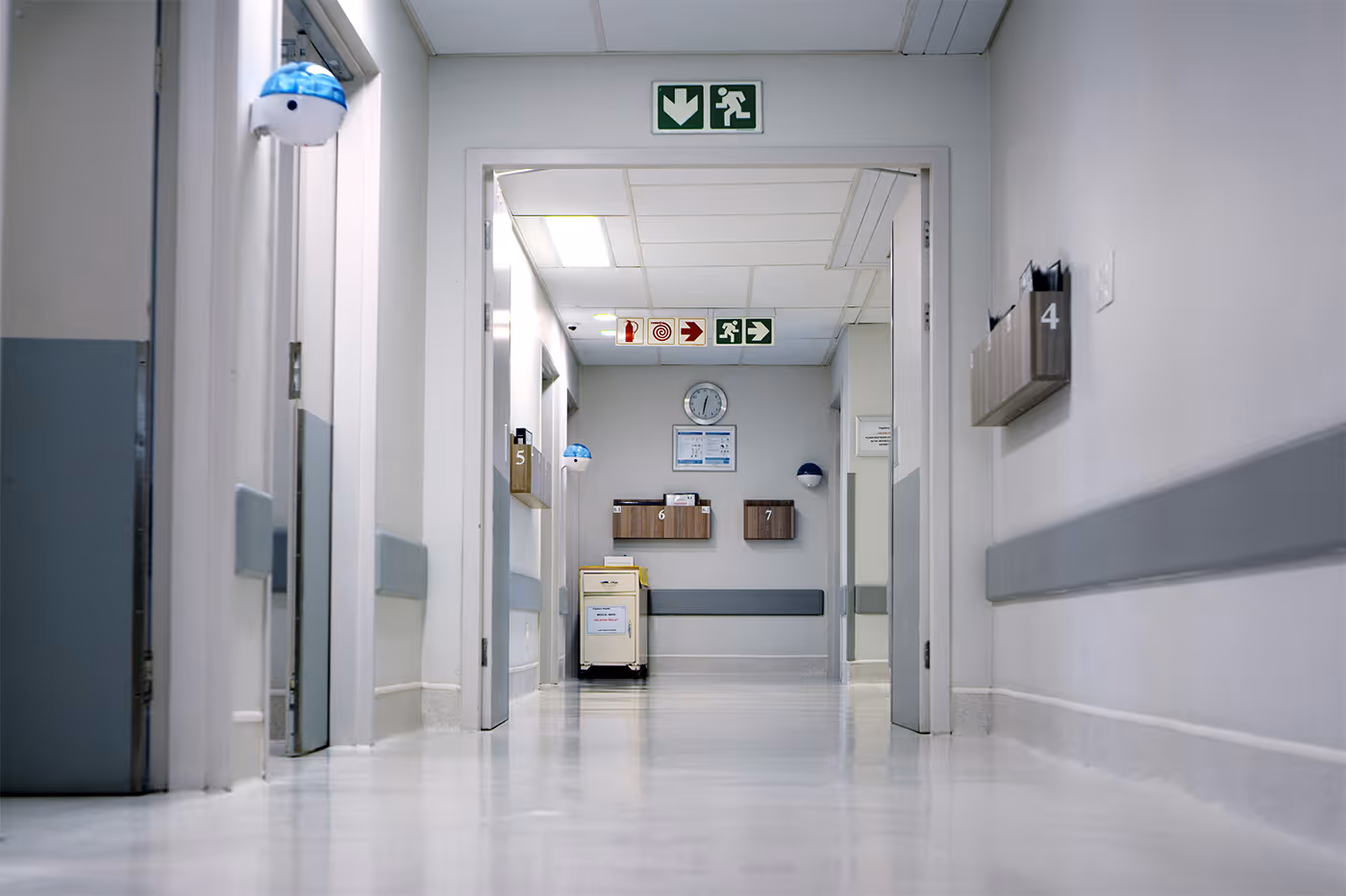Empty hospital corridor with white walls, open doors, and emergency exit signs overhead.