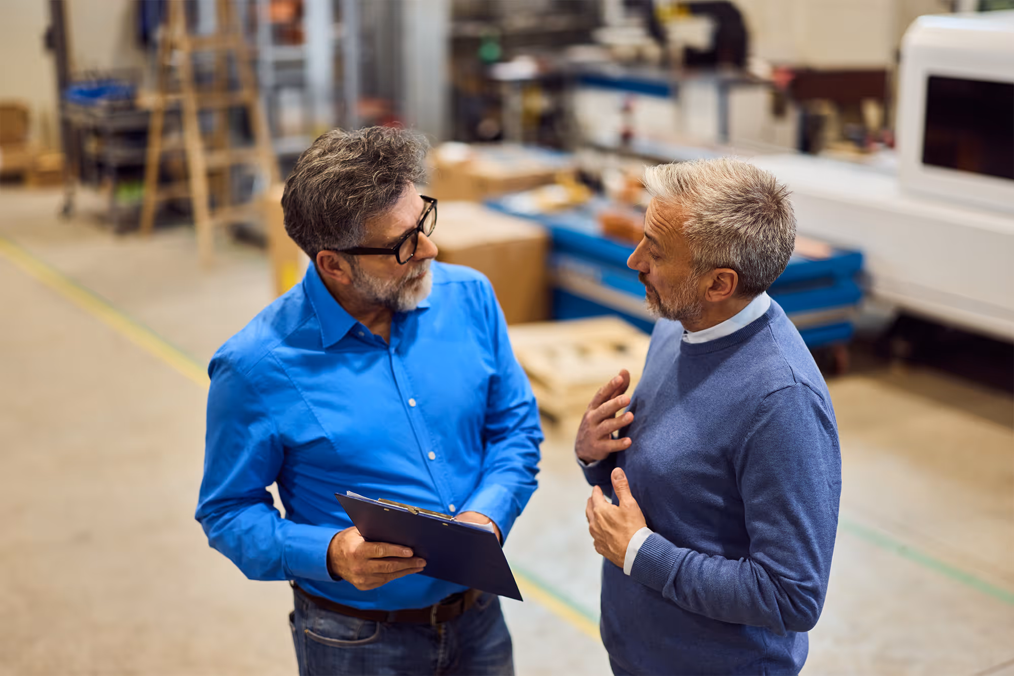 Two men in a factory having a discussion; one holds a clipboard, the other gestures with his hands.