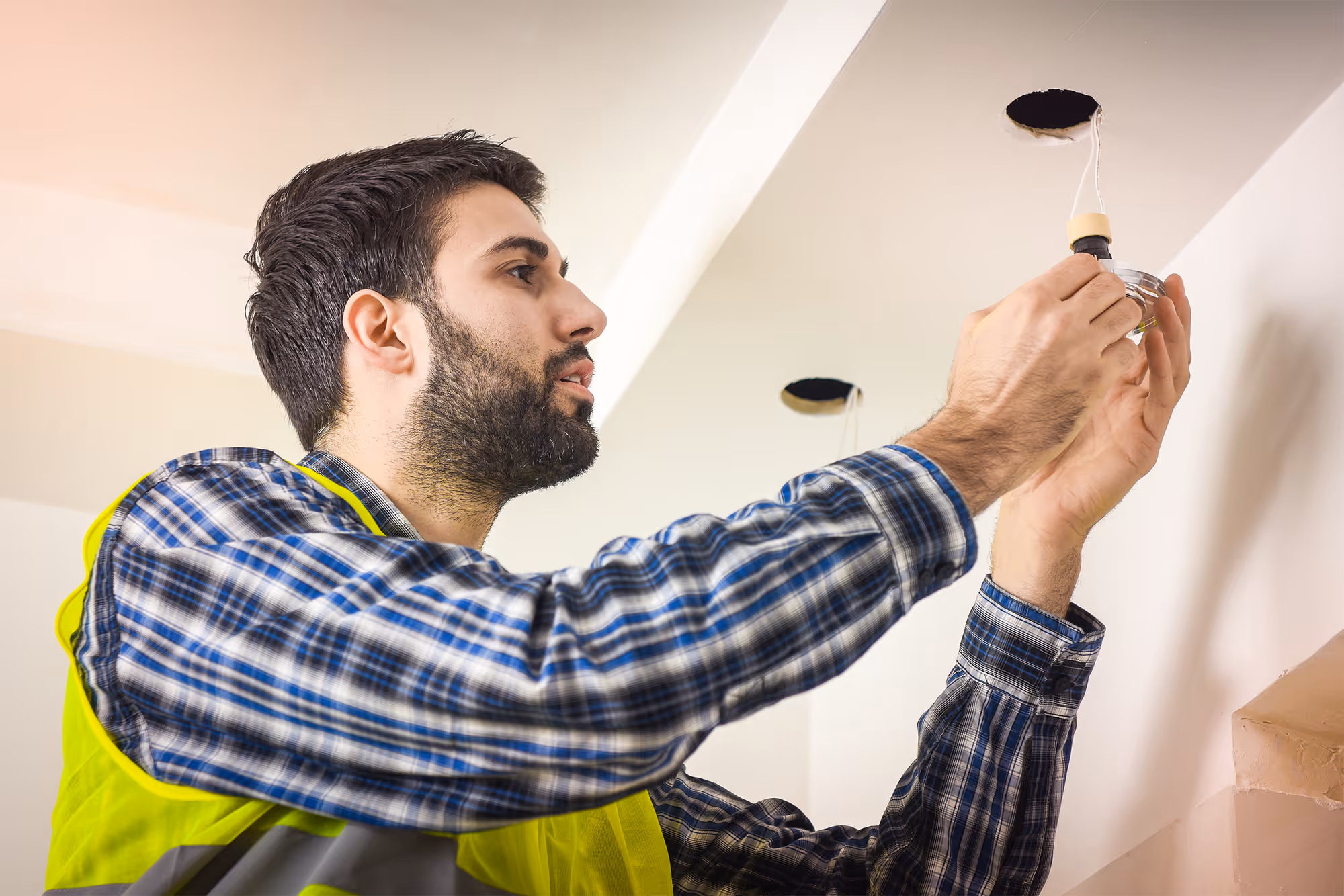 Man wearing a safety vest installing a recessed light fixture in a ceiling hole indoors.