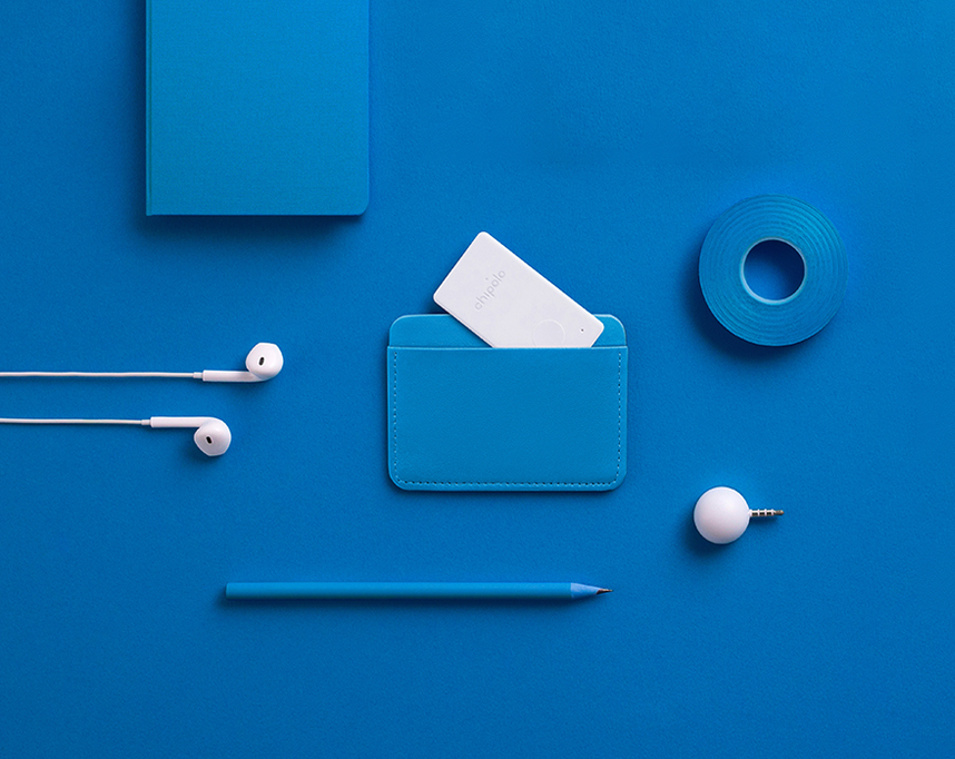 Flat lay of blue office supplies including a notebook, pencil, adhesive tape, white earphones, a blue card wallet with a white Chipolo tracker, and a white spherical auxiliary jack.