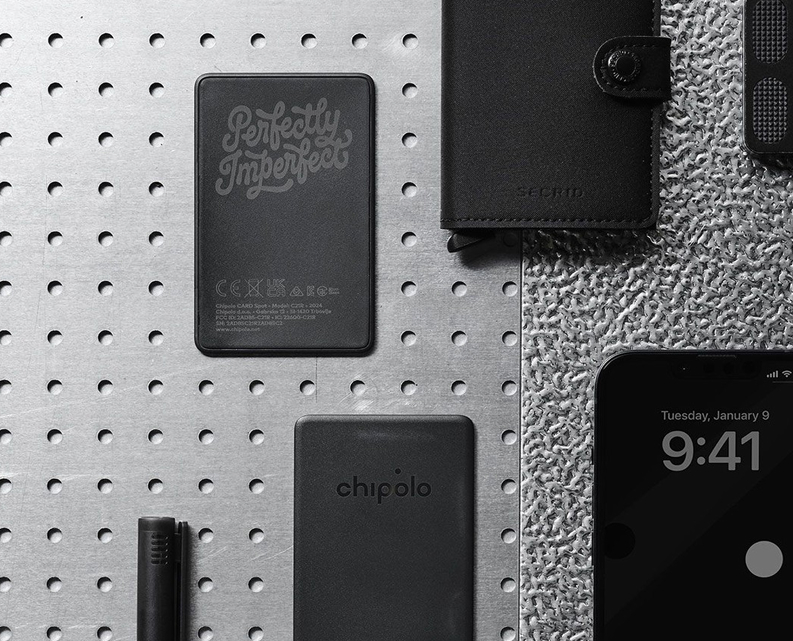 Flat lay of black tech accessories including a Chipolo tracker, a wallet with a snap button, and a smartphone showing 9:41 on Tuesday, January 9, all on a perforated metal surface.