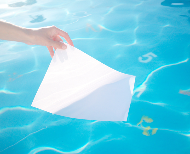 A hand holding a blank white piece of paper partially submerged in clear blue pool water with depth markings visible.