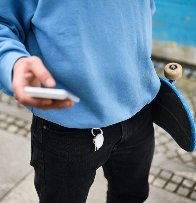 Person in blue sweatshirt holding a smartphone with a skateboard tucked under arm and a white key tracker attached to black pants.