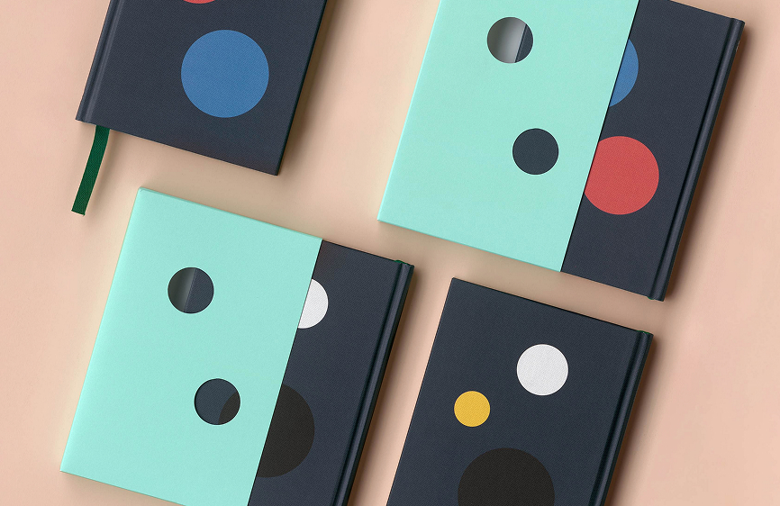 Four closed notebooks with dark covers and colorful circles, two with light blue slipcovers featuring cut-out circles, arranged on a beige surface.