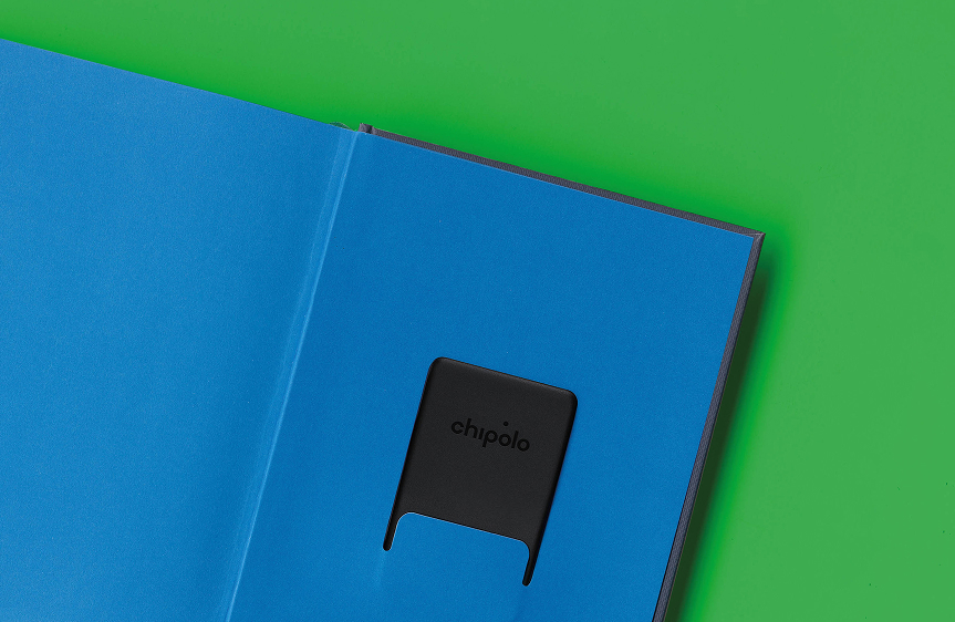 Black Chipolo tracking device placed in a blue slot of an open notebook on a green surface.