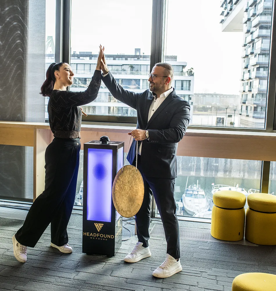 Zwei Personen geben sich ein High Five vor einem Fenster, neben ihnen ein Gong und eine Säule.