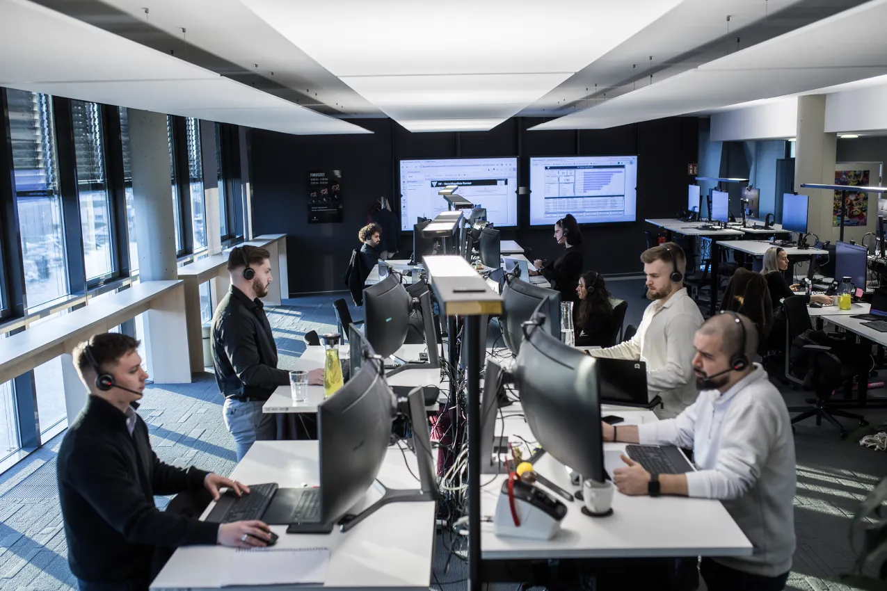 Büro mit mehreren Mitarbeitenden an Computern mit Headsets, beleuchtet von Tageslicht durch Fenster.